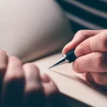 uma mão feminina segura uma caneta sobre um caderno em branco, pronta para escrever.