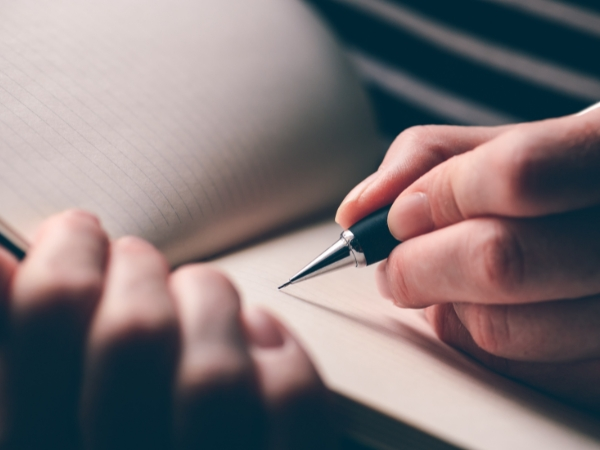 uma mão feminina segura uma caneta sobre um caderno em branco, pronta para escrever.